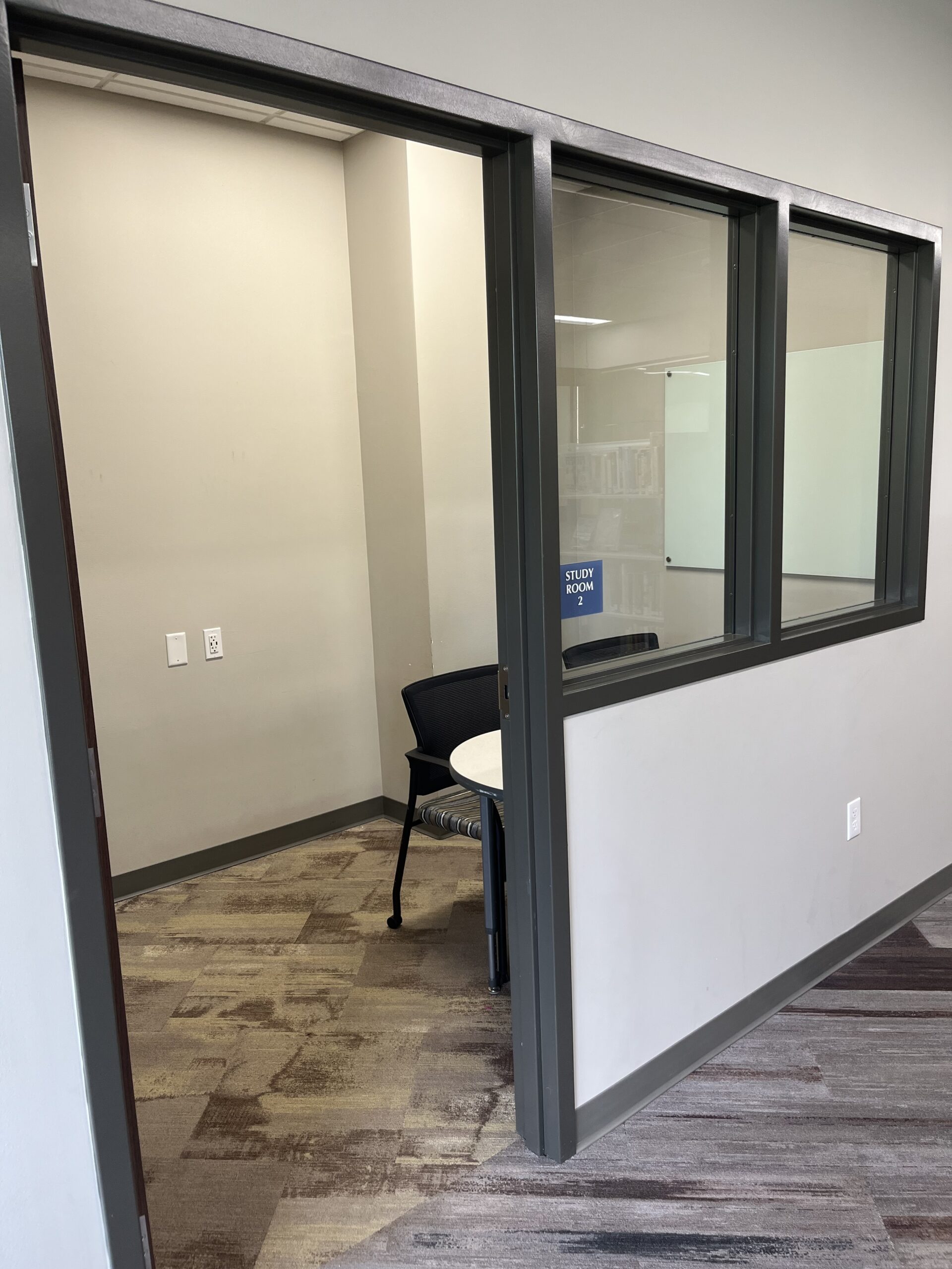 Small, glass-walled study room with a table and two chairs on a patterned carpet. Soft lighting and a whiteboard add a calm, focused atmosphere.