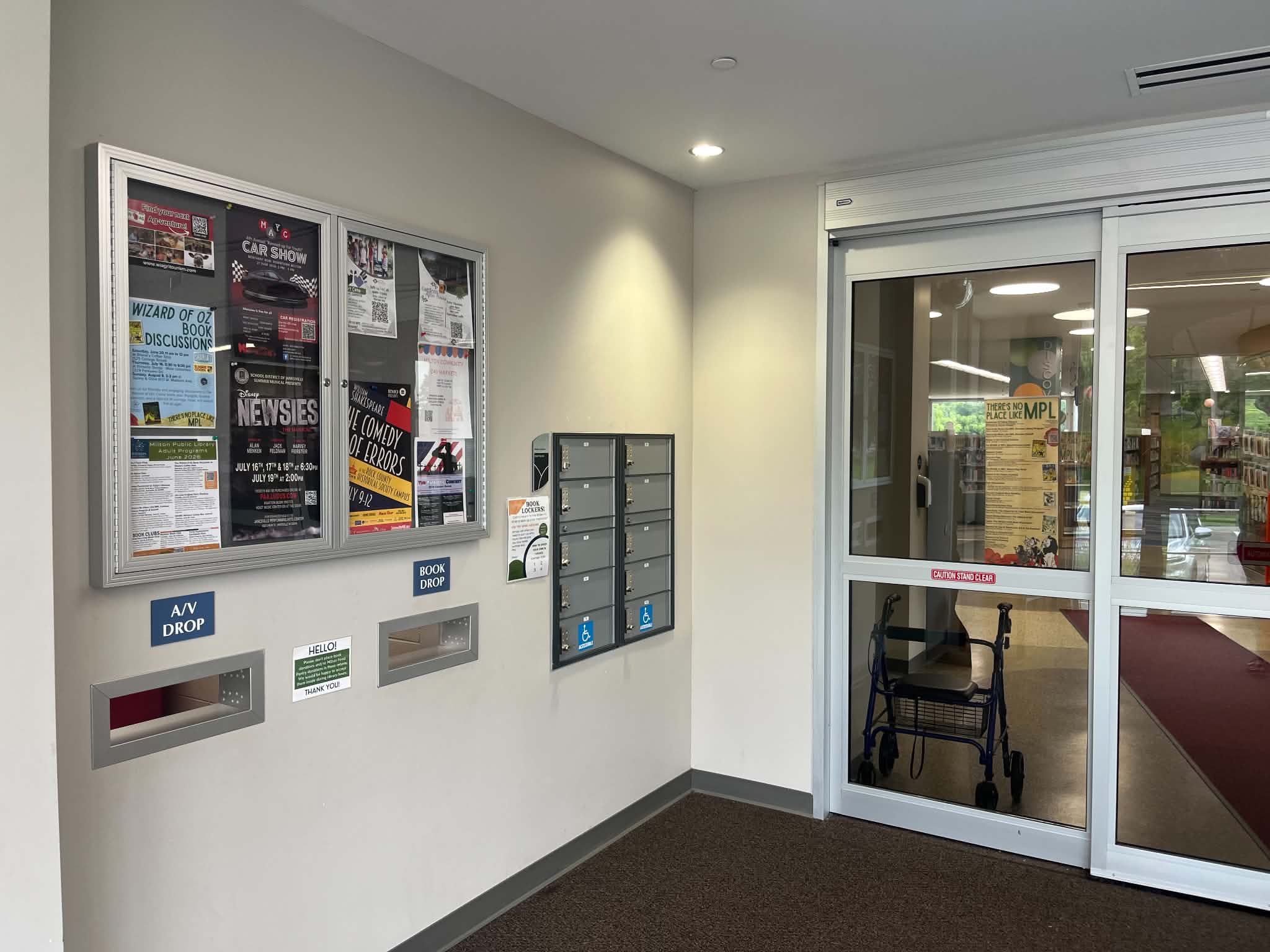 A building lobby with bulletin boards, mailboxes, book and AV drop slots, and a walker near automatic glass doors leading to an interior space.