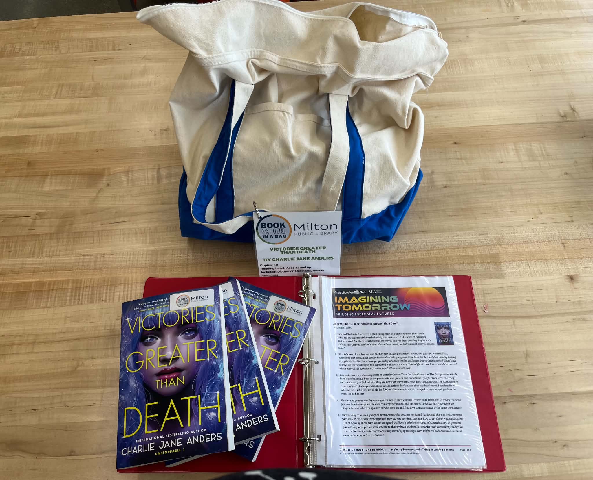A tote bag labeled "Milton Public Library" on a table with books titled "Victories Greater Than Death" and an open binder. Cozy and engaging