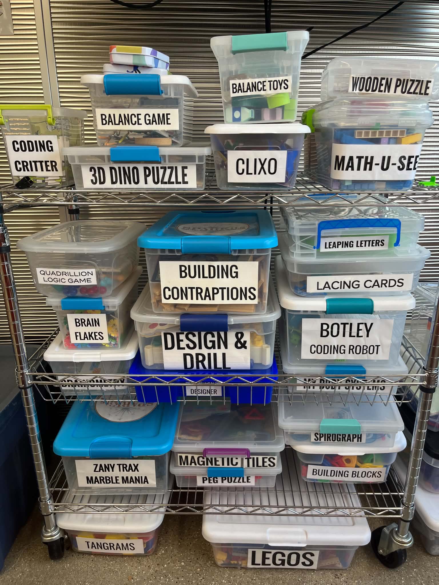 A metal shelving unit holds various labeled plastic containers with educational toys and puzzles, organized into three rows.<br />
