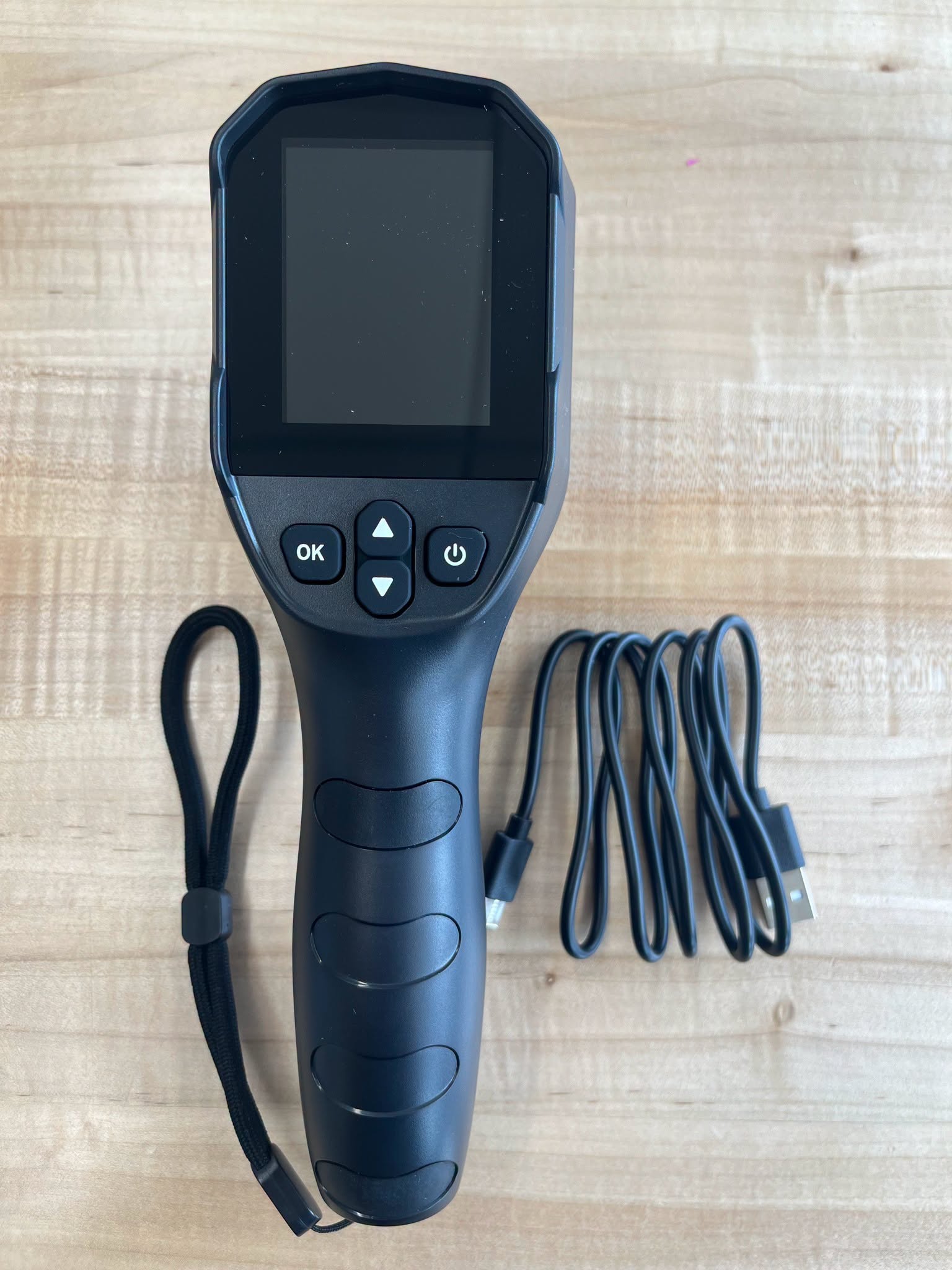A black handheld thermal imaging camera with a screen, navigation buttons, and a power button, accompanied by a black USB charging cable on wood.<br />

