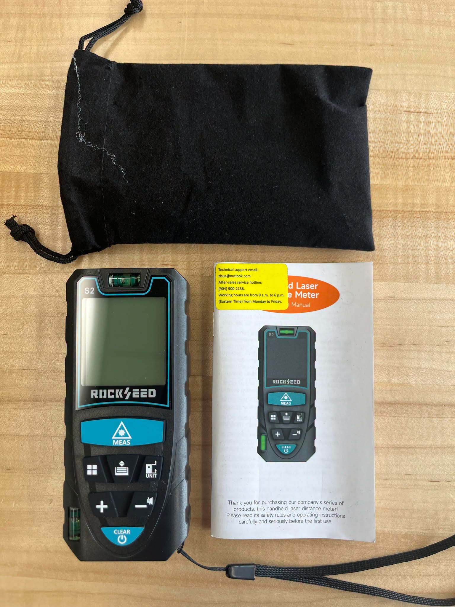 A black laser distance meter, its user manual, and a black drawstring storage pouch are arranged on a light wood grain surface.<br />
