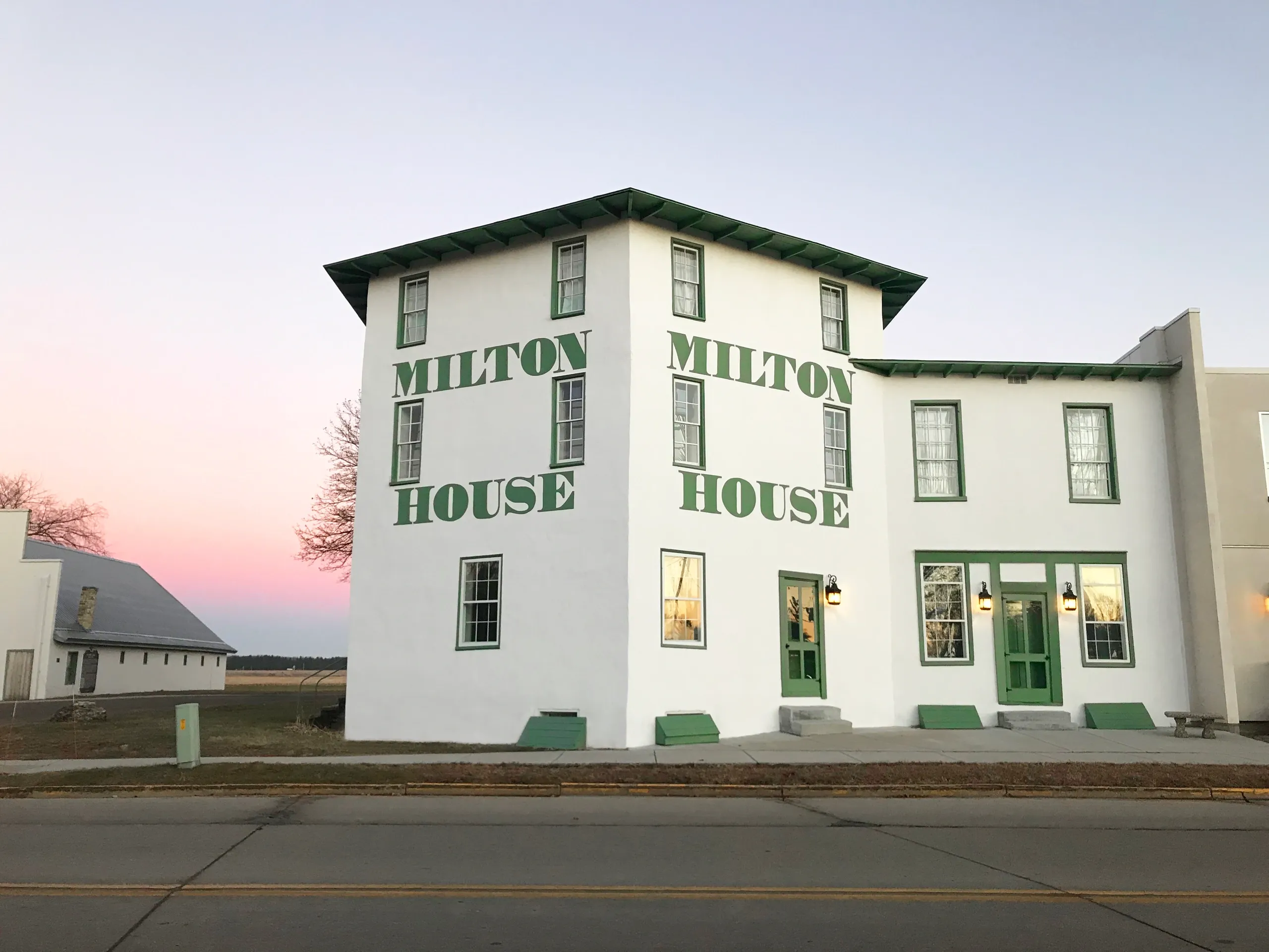 A white three-story building with green trim and large “Milton House” text on the exterior, under a clear sky at dusk.