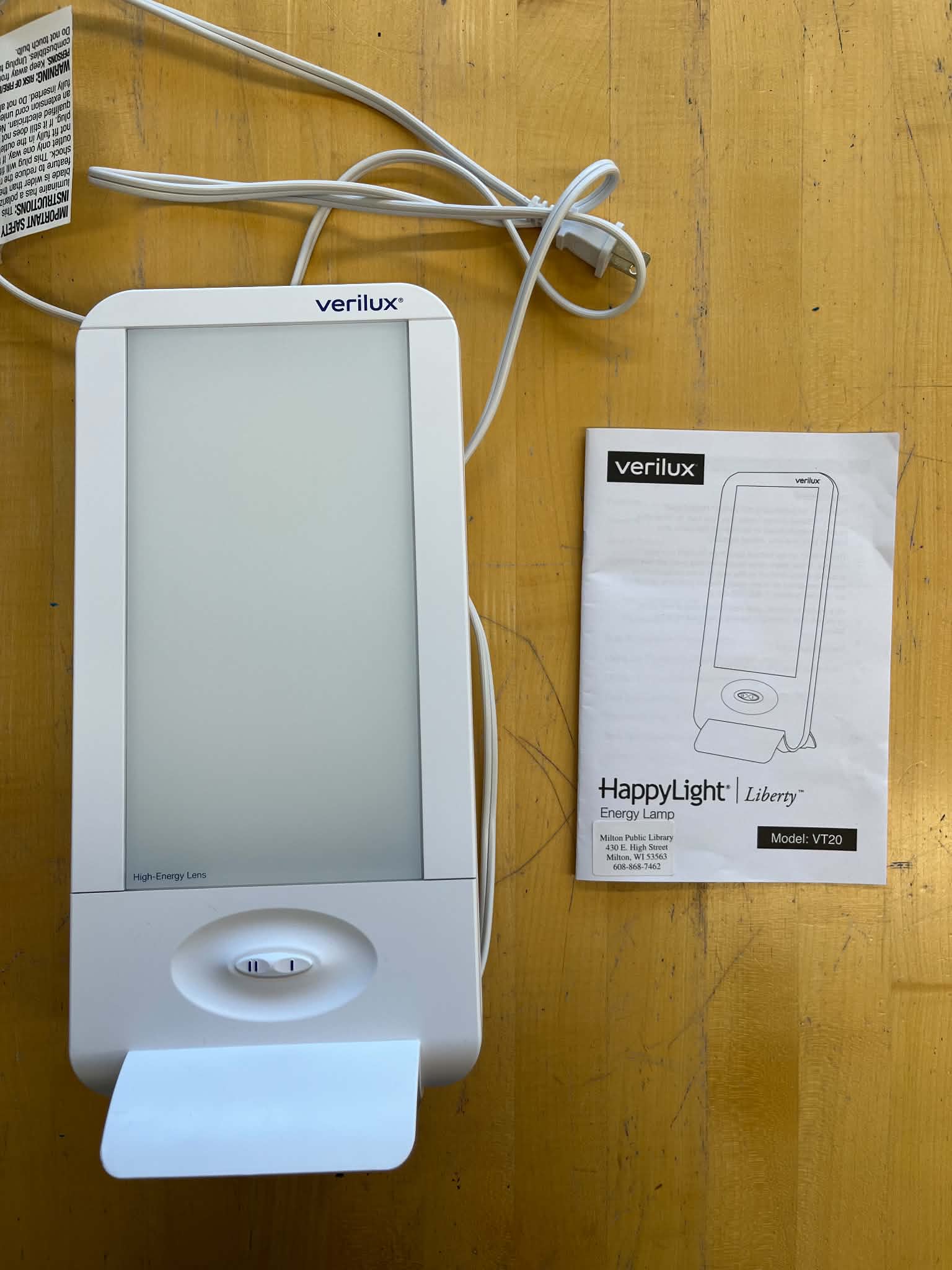 A white Verilux HappyLight Liberty energy lamp with a power cord and its instruction manual placed on a light-colored wooden surface.<br />
