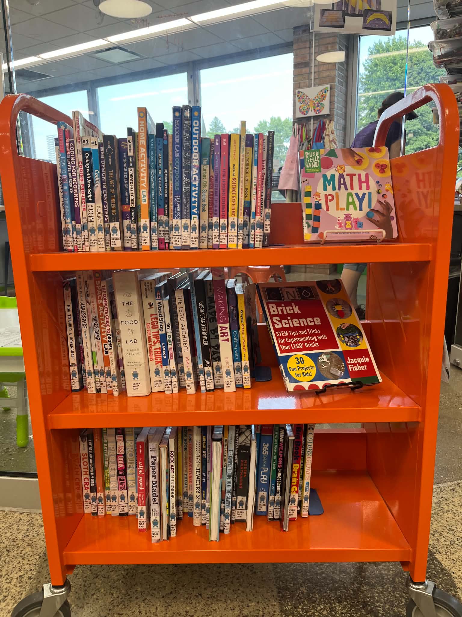 An orange book cart filled with colorful children's books, including a book titled "MATH PLAY!" in a well-lit library setting.<br />
