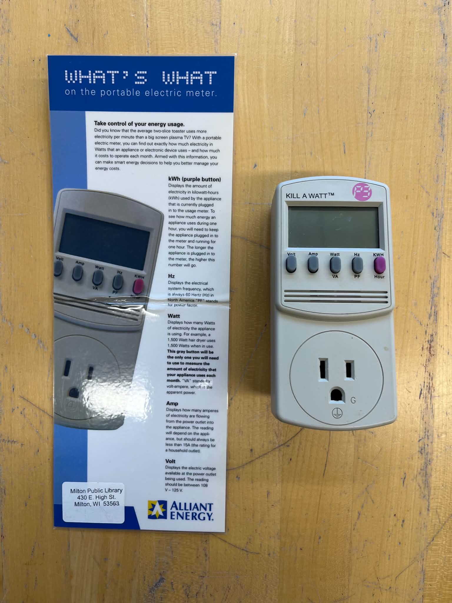 A Kill A Watt electricity usage monitor sits next to its instructional pamphlet on a wooden surface, detailing how to measure appliance energy use.<br />
